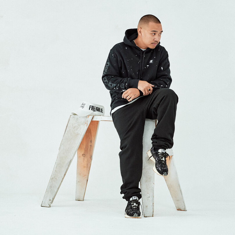 Man sitting on a saw horse in a grey studio environment wearing pieces from the Joshua Vides collection.