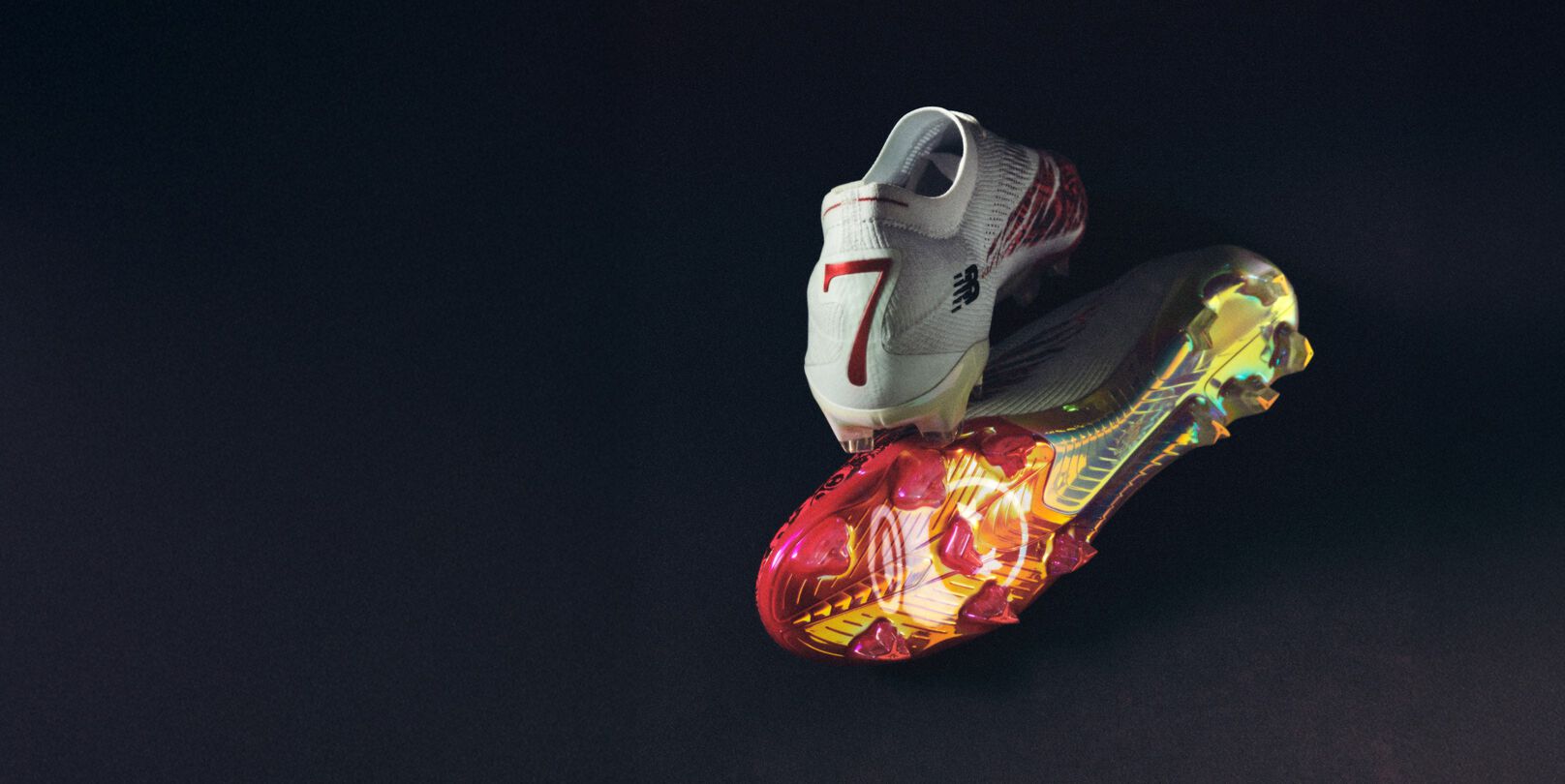 Pair of white soccer cleats with red accents and multicolor sole on a dark background. 
