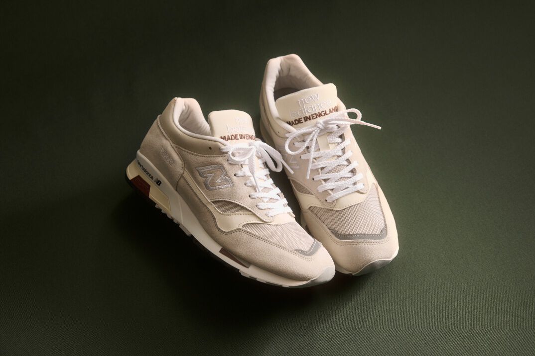 Knee down view of a man wearing a pair of New Balance Made in UK sneakers standing on a rustic wooden floor with smashed blue and white ceramic crockery all over the ground and white cabinets in the background. 