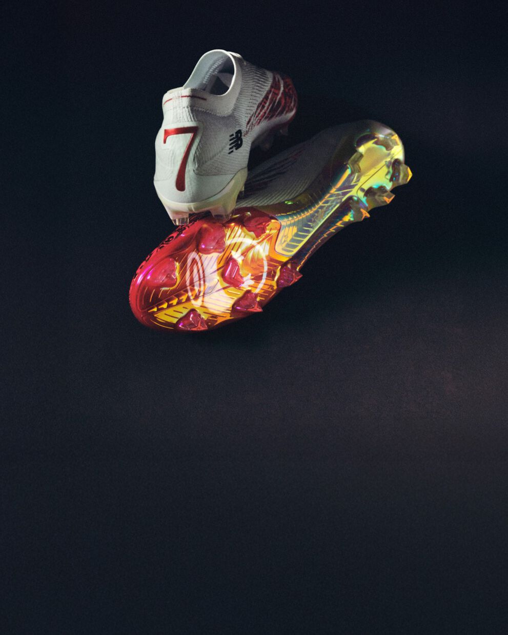 Pair of white soccer cleats with red accents and multicolor sole on a dark background. 